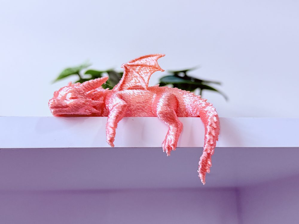Articulated baby dinosaur figure printed in silk copper PLA, curled up on a shelf